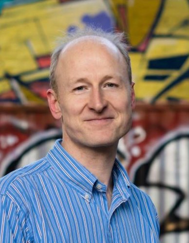 Peter McGeeney, smiling, in a blue striped shirt in front of a graffiti wall